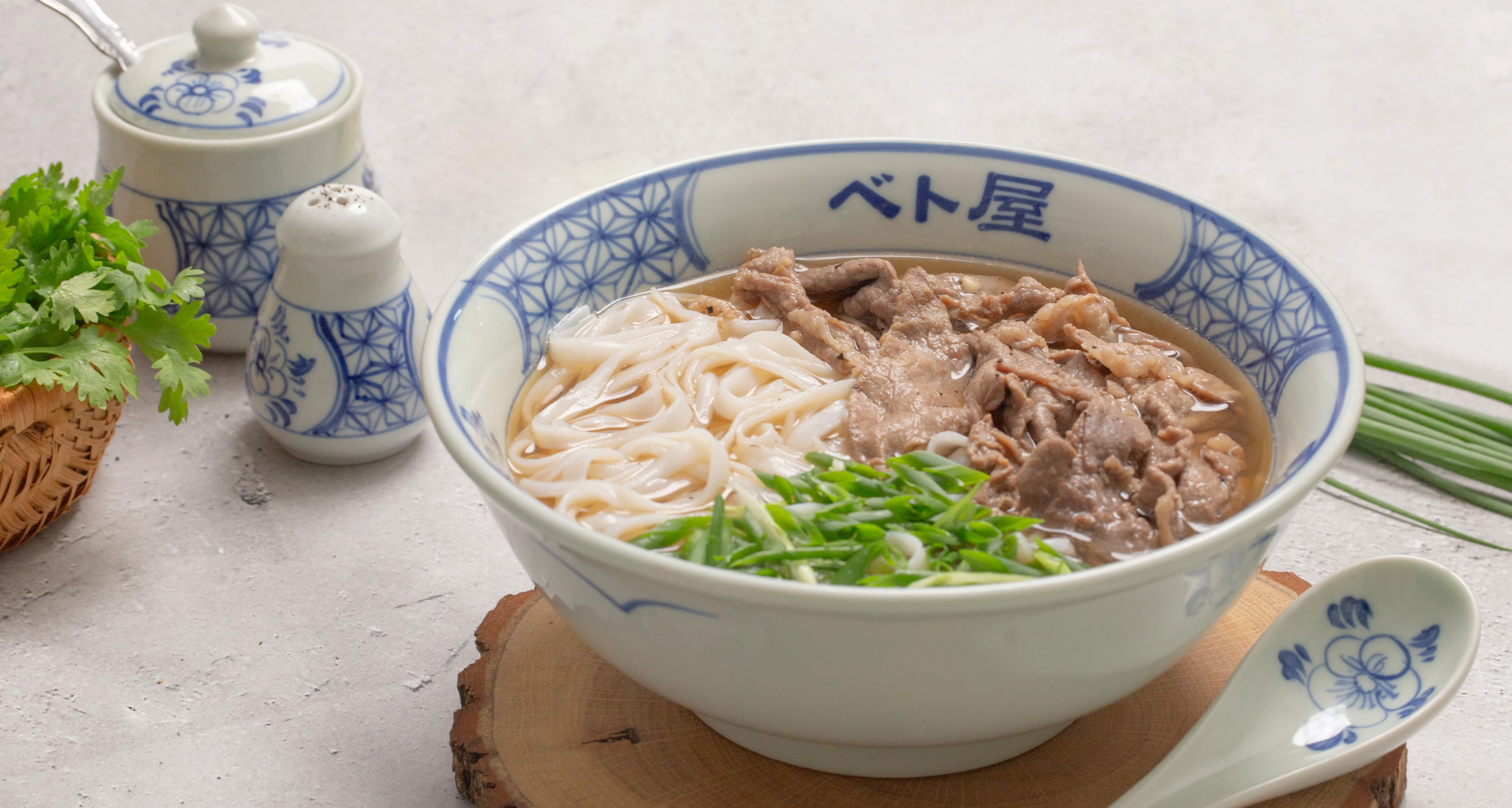 Bát phở bò Viet Origin đặt trên khoanh gỗ, bên cạnh là giỏ rau mùi và hũ gia vị