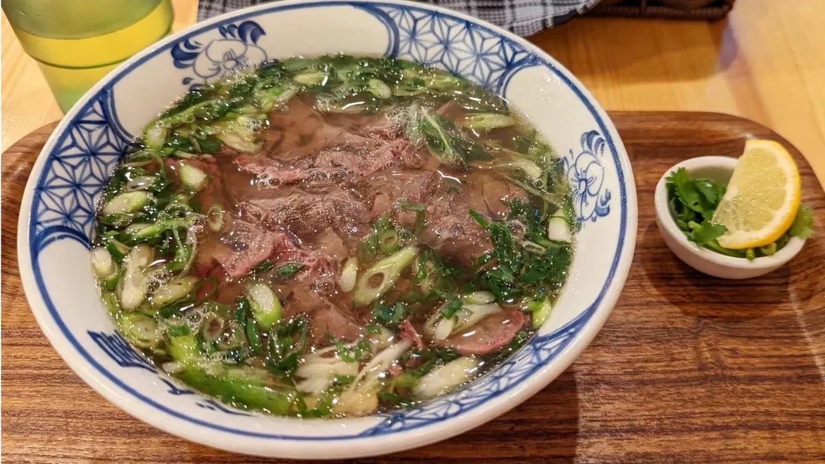 木のトレーにのった、青ねぎとパクチーをたっぷりのせた牛肉のフォーと、レモンとパクチーの小皿、冷たいお茶のグラス