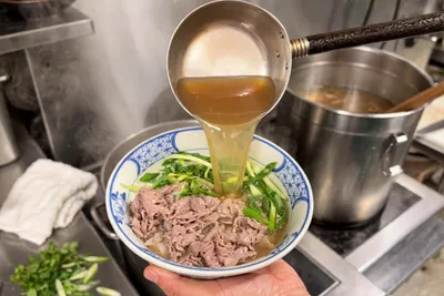 湯気の立つ厨房で、手に持った丼の牛肉と青ねぎの上に、お玉から澄んだ黄金色のスープを注ぐ瞬間